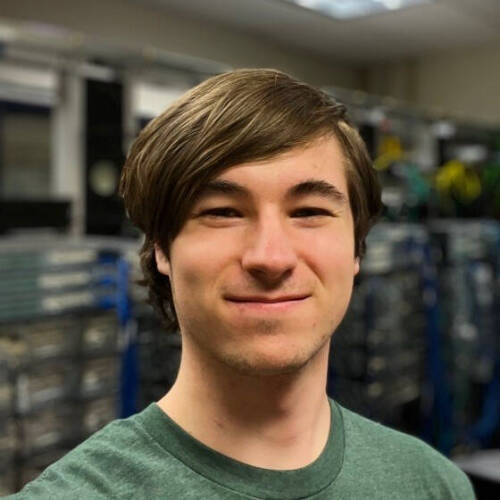Logan Nommensen, wearing a green T-shirt smiles at the camera. In the background there's a blurred image of a server room.